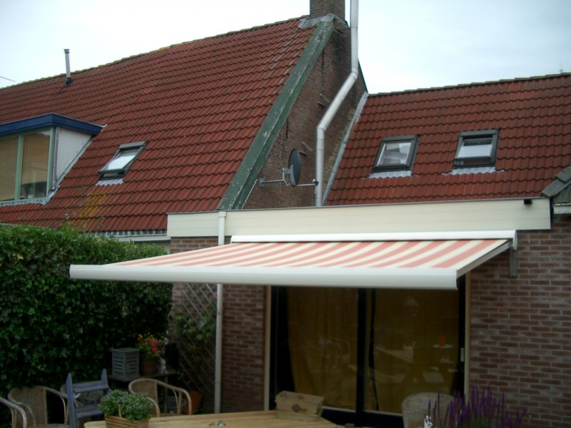 Wit zonnescherm met rode strepen boven een tuindeur aan een huis met dakkapellen