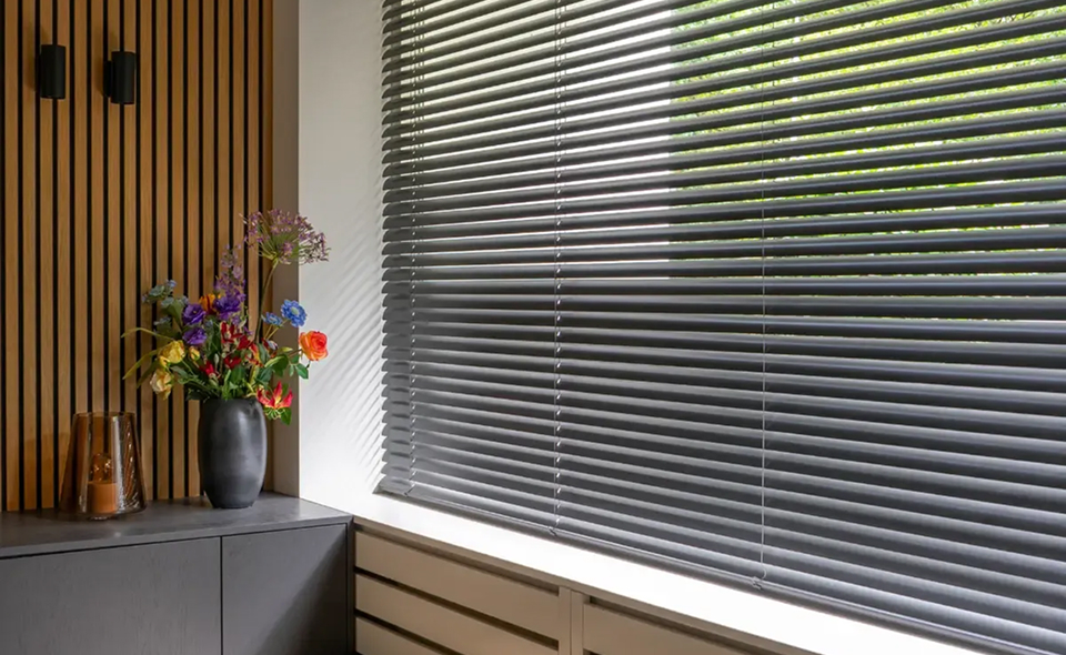 Grijze horizontale jaloezieën voor een raam met bloemenvaas en decoratieve lamp ervoor