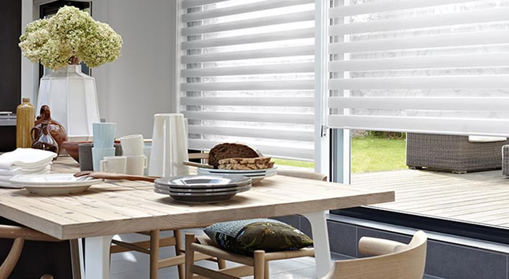 Houten eettafel met serviesgoed en vaas met bloemen voor raam met horizontale rolgordijnen
