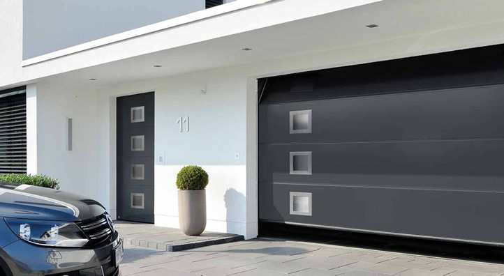 Automatische garagedeur in zwart met sensoren en een auto geparkeerd voor een moderne woning