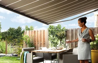 Vrouw onder buitenzonwering die een glas water inschenkt bij een tuintafel met tuinmeubilair en planten
