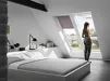 Vrouw staat bij VELUX dakraam in slaapkamer met open rolgordijn